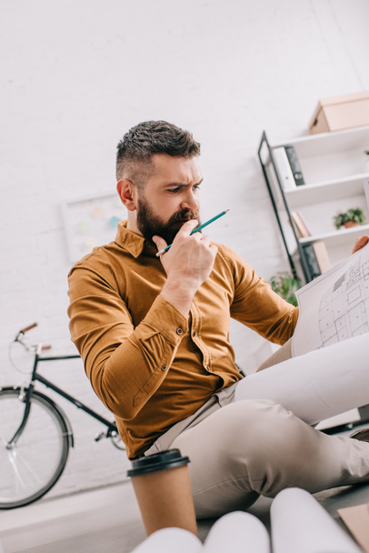 задумчивый бородатый взрослый мужчина-архитектор с чертежом и работающий над проектом в офисе
  - Фото, изображение