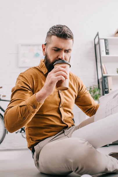 Porträt eines bärtigen erwachsenen männlichen Architekten, der Coffee to go trinkt und im Büro an einem Entwurf arbeitet  - Foto, Bild
