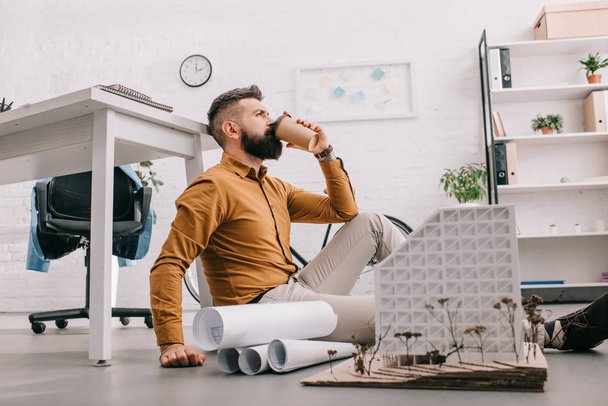 bearded adult male architect drinking coffee to go and working on construction project with blueprints in office  - Photo, Image