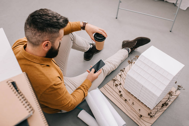 Blickwinkel des bärtigen erwachsenen männlichen Architekten mit Smartphone, Kaffee to go in der Hand und Arbeit an Entwürfen im Büro  - Foto, Bild