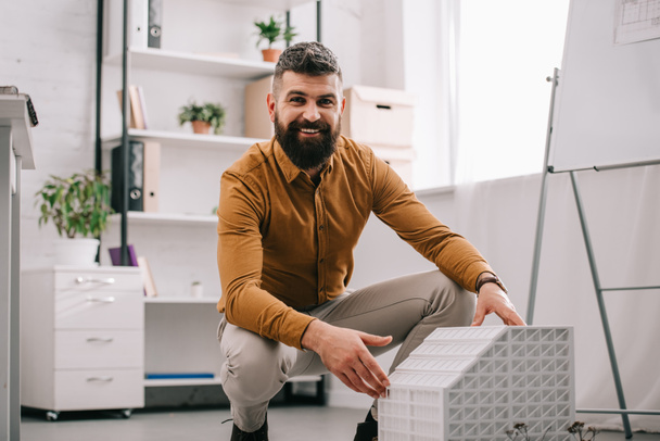 smiling bearded adult male architect looking at camera and presenting house model in office  - Foto, Imagen