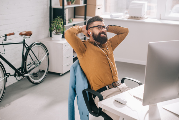smiling bearded adult businessman in earphones with hands behind head sitting and relaxing at computer desk in office - 写真・画像