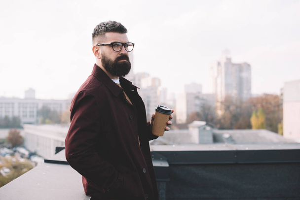 focused stylish adult man in glasses holding coffee to go on rooftop - Photo, Image