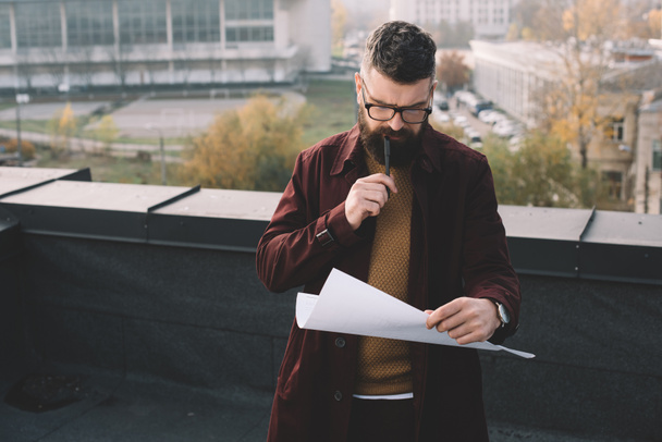 pensive adult male architect in glasses holding blueprint and working on project on rooftop - Фото, изображение