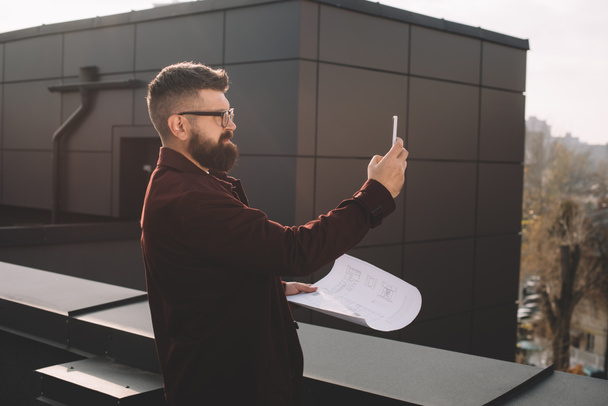 serious adult male architect in glasses taking photo and holding blueprint on rooftop - Фото, изображение