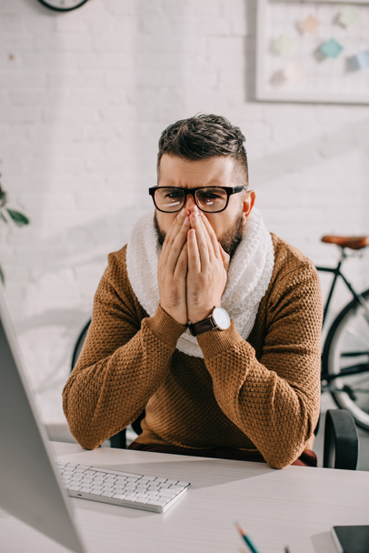nemocný podnikatel v pletená šála sedí v kanceláři, kašel a pokrývající ústa rukama - Fotografie, Obrázek