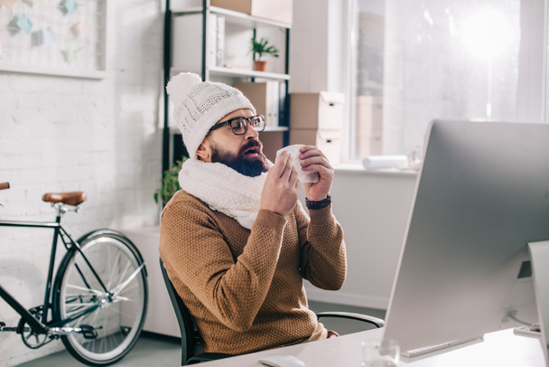 hombre de negocios enfermo en bufanda y sombrero de punto sentado en el escritorio de la oficina y estornudos
  - Foto, Imagen