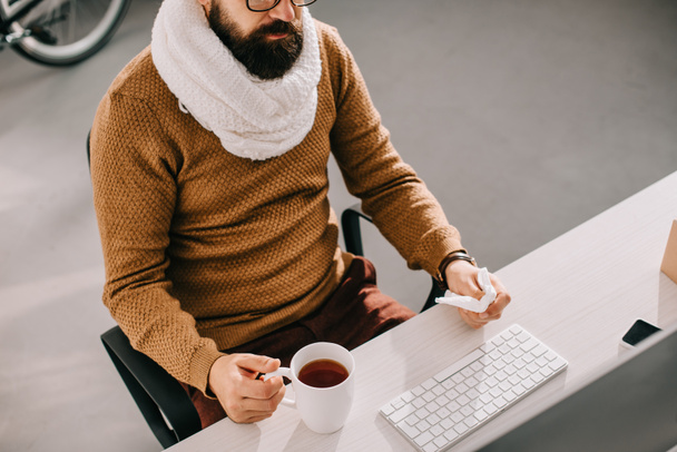 sick businessman in scarf holding cup of tea and tissue and sitting at office desk - Foto, Imagem