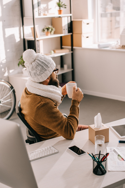 хворий бізнесмен в шарфі і в'язаний капелюх сидить за офісним столом і тримає чашку чаю
  - Фото, зображення