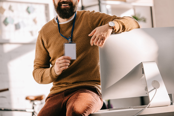 обрізаний вид бородатого дорослого бізнесмена, що сидить на столі і тримає порожню мітку безпеки в офісі
 - Фото, зображення