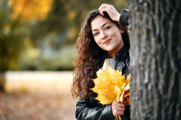 Γυναίκα με τα κίτρινα φύλλα στέκονται κοντά μεγάλο δέντρο στο Φθινοπωρινό πάρκο της πόλης. Πορτρέτο κινηματογράφηση σε πρώτο πλάνο. - Φωτογραφία, εικόνα