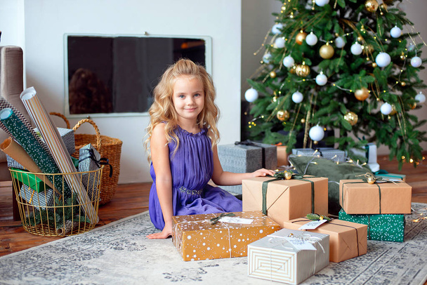A little blond girl in colorful dress sits in the room on backround gifts. Holiday concept. Girl on photo. Merry Cristmas, happy holiday - Foto, imagen