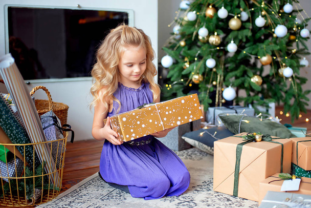 A little blond girl in colorful dress sits in the room on backround gifts. Holiday concept. Girl on photo. Merry Cristmas, happy holiday - 写真・画像