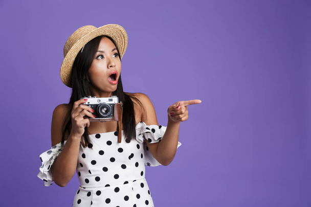 Portrait of a cheerful asian woman in summer hat standing isolated over violet background, holding photo camera, pointing away - Photo, Image