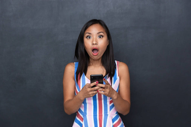 Immagine di donna asiatica sorpresa 30s utilizzando il telefono cellulare nero in piedi isolato su sfondo grigio
 - Foto, immagini