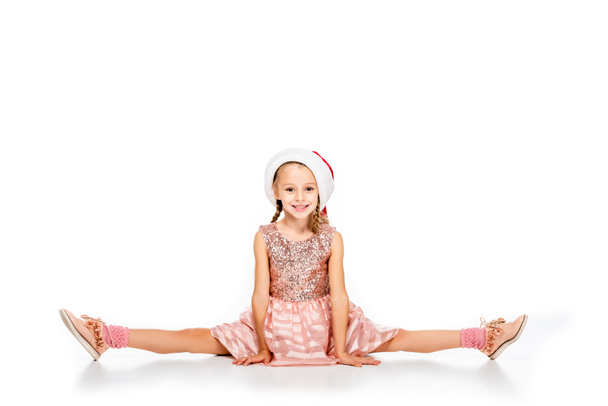 happy little child in santa hat sitting splits on floor and looking at camera isolated on white - Photo, Image