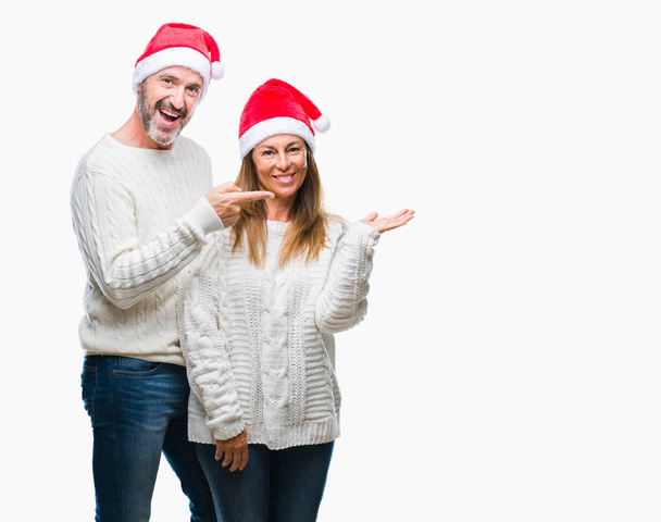Латиноамериканская пара средних лет в рождественской шляпе на изолированном фоне удивлена и улыбается в камеру, представляя с рукой и указывая пальцем
. - Фото, изображение