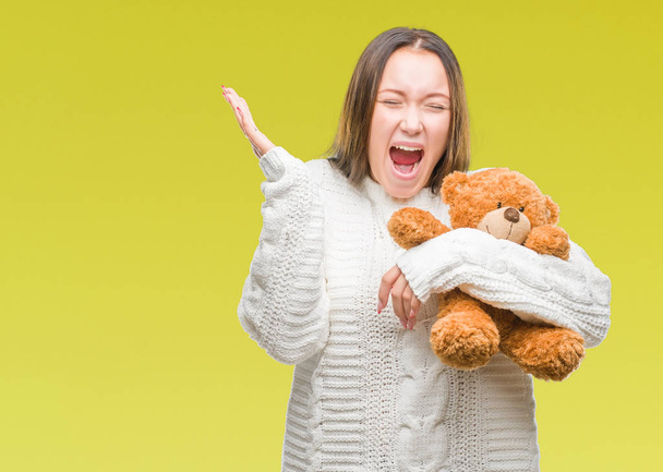 Young caucasian woman holding teddy bear over isolated background very happy and excited, winner expression celebrating victory screaming with big smile and raised hands - 写真・画像