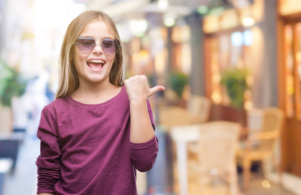 Young beautiful girl wearing sunglasses over isolated background smiling with happy face looking and pointing to the side with thumb up. - Fotografie, Obrázek