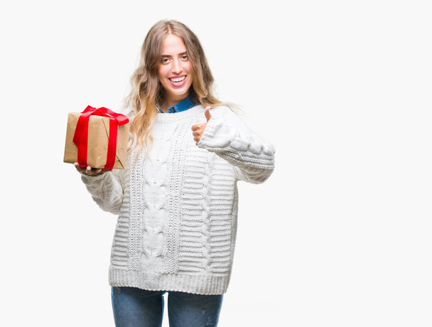 Beautiful young blonde woman holding gift over isolated background happy with big smile doing ok sign, thumb up with fingers, excellent sign - Photo, Image