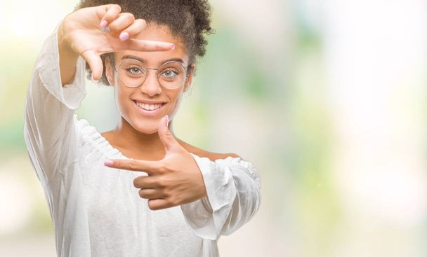 Mujer afroamericana joven con gafas sobre fondo aislado sonriendo haciendo montura con manos y dedos con cara feliz. Concepto de creatividad y fotografía
. - Foto, Imagen