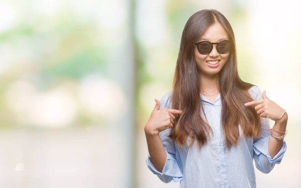 Fiatal ázsiai nő napszemüveget visel, mint elszigetelt háttér látszó magabiztos mosollyal arcán, mutatva magát, ujjaival, büszke és boldog. - Fotó, kép