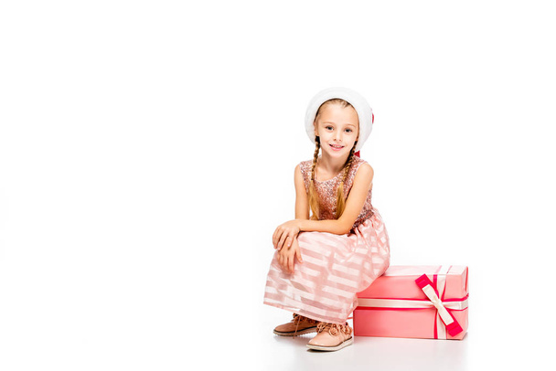 adorable little child in santa hat sitting on gift box and looking at camera isolated on white - Photo, Image