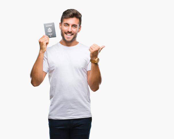 Jonge knappe man met paspoort van de Verenigde Staten over geïsoleerde achtergrond aan te wijzen en te tonen met de duim aan de kant met een blij gezicht lachend - Foto, afbeelding
