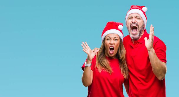 中年ヒスパニック系カップルのクリスマス帽子をかぶっている狂気の背景を分離し、怒って叫んで、叫んで積極的な表現と腕を上げます。欲求不満のコンセプト. - 写真・画像