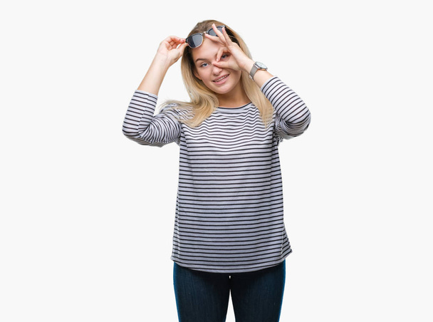 Young caucasian woman wearing sunglasses over isolated background doing ok gesture with hand smiling, eye looking through fingers with happy face. - Foto, Bild
