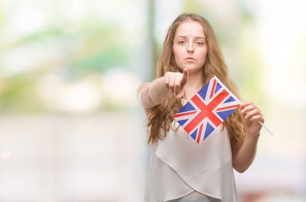 Giovane donna bionda che tiene la bandiera del Regno Unito puntando con il dito alla fotocamera e a te, segno della mano, gesto positivo e fiducioso dalla parte anteriore
 - Foto, immagini