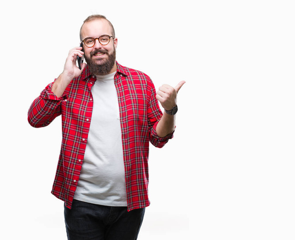Young caucasian hipster man talking on smartphone over isolated background pointing and showing with thumb up to the side with happy face smiling - Photo, Image