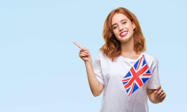 Молодая красивая женщина с флагом Соединенного Королевства на изолированном фоне очень счастлива указывая рукой и пальцем в сторону
 - Фото, изображение
