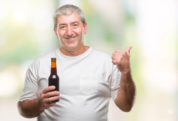 Guapo hombre mayor bebiendo botella de cerveza sobre fondo aislado apuntando y mostrando con el pulgar hacia el lado con la cara feliz sonriendo
 - Foto, imagen