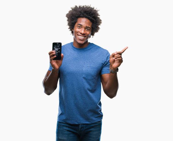 Afroamerikaner hält kaputtes Smartphone über isoliertem Hintergrund und zeigt glücklich mit Hand und Finger zur Seite - Foto, Bild