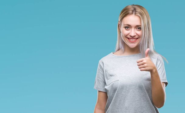 Young blonde woman over isolated background doing happy thumbs up gesture with hand. Approving expression looking at the camera with showing success. - Photo, Image