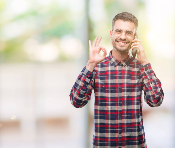 Young hipster adult man talking on the phone doing ok sign with fingers, excellent symbol - Fotó, kép