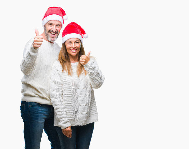 Średnim wieku hiszpanin para sobie Boże Narodzenie kapelusz na białym tle robi szczęśliwy kciuk w górę gest ręką. Zatwierdzająca wyrażenie patrząc na kamery z pokazując sukces. - Zdjęcie, obraz