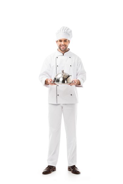 pohledný mladý kuchař drží tác s kupolí a při pohledu na fotoaparát izolované na bílém - Fotografie, Obrázek