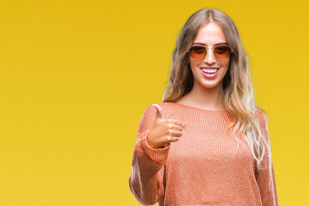 Beautiful young blonde woman wearing sunglasses over isolated background doing happy thumbs up gesture with hand. Approving expression looking at the camera with showing success. - Photo, Image