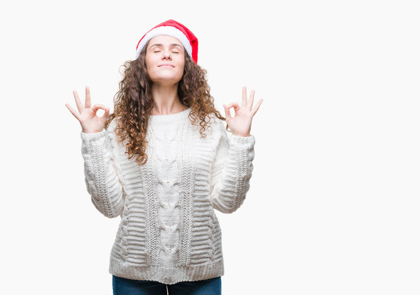 孤立した背景の上身に着けているクリスマスの帽子を若いブルネットの少女リラックスして瞑想の指ジェスチャーをやって閉じた目と笑顔します。ヨガの概念. - 写真・画像