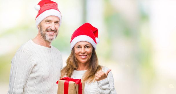 hispanisches Paar mittleren Alters mit Weihnachtsmütze und Geschenk über isoliertem Hintergrund zeigend und mit Daumen zur Seite zeigend mit glücklichem Lächeln im Gesicht - Foto, Bild
