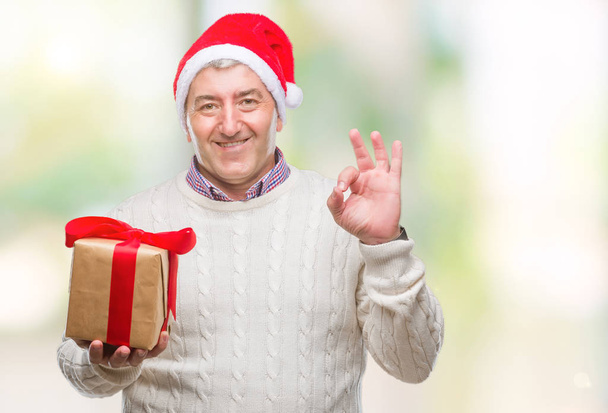 Komea vanhempi mies yllään joulu hattu ja tilalla lahja yli eristetty tausta tekee ok merkki sormilla, erinomainen symboli
 - Valokuva, kuva