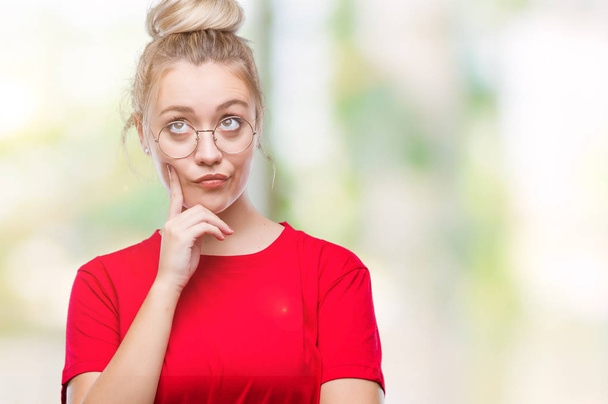 Young blonde woman wearing glasses over isolated background with hand on chin thinking about question, pensive expression. Smiling with thoughtful face. Doubt concept. - Photo, image