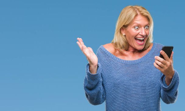 Middle age blonde woman sending message using smartphone over isolated background very happy and excited, winner expression celebrating victory screaming with big smile and raised hands - Photo, Image