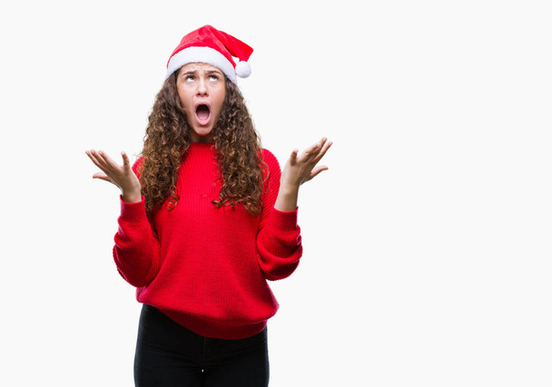 Молода брюнетка в різдвяному капелюсі над ізольованим фоном божевільний і божевільний крик і крик з агресивним виразом і піднятими руками. Концепція розмальовки
. - Фото, зображення