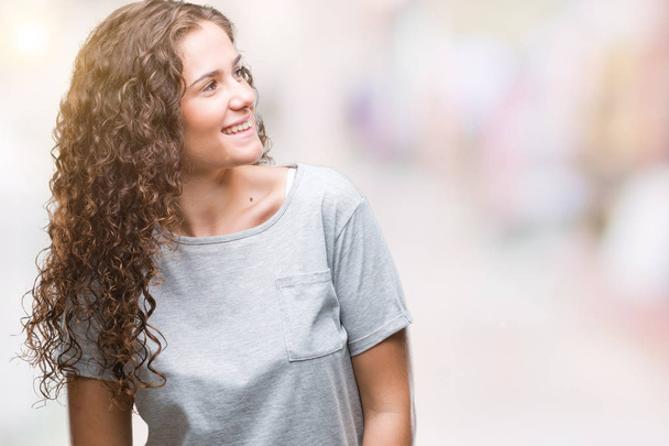Beautiful young brunette curly hair girl wearing casual look over isolated background looking away to side with smile on face, natural expression. Laughing confident. - Photo, Image