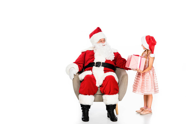 Санта Клаус сидит в кресле и дарит подарок маленькому ребенку, изолированному на белом
 - Фото, изображение