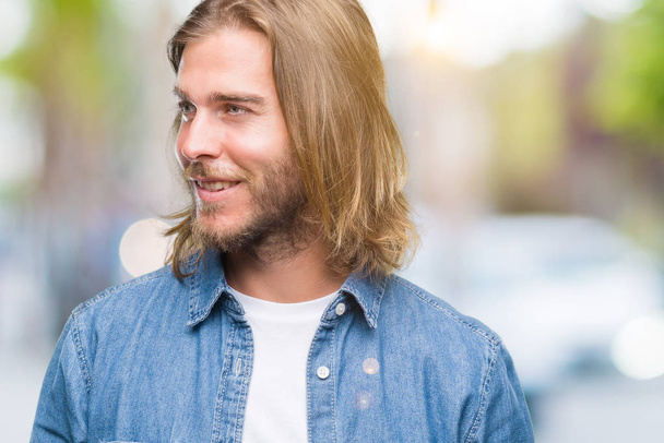 Young handsome man with long hair over isolated background looking away to side with smile on face, natural expression. Laughing confident. - Photo, Image