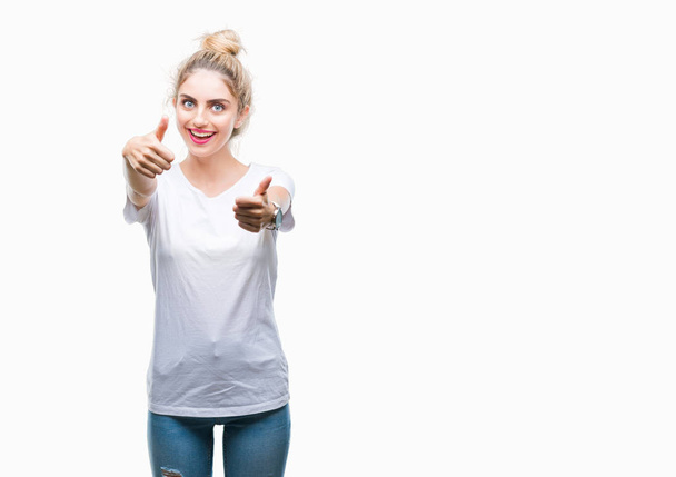 Молодая красивая блондинка в белой футболке на изолированном фоне, одобряющая позитивный жест рукой, улыбается и радуется успеху. Глядя в камеру, жест победителя
. - Фото, изображение